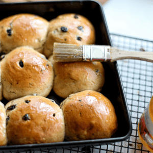 A tray of freshly baked currant buns being brushed with apricot jam.